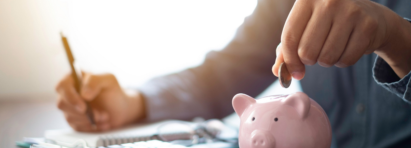Ein Mensch sitz am Schreibtisch. Auf dem Schreibtisch liegen Unterlagen, ein Taschenrechner und ein Sparschwein. Mit der rechten Hand schreibt die Person in den Unterlagen und mit der rechten Hand wirft sie eine Münze in das Sparschwein.