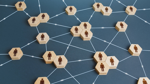 Auf einer grauen Fläche liegen verschiedene sechseckige Holztäfelchen. Auf den einzelnen Holztäfelchen ist eine schematische Abbildung einer Person gedruckt. Die Holztäfelchen sind mit Linien verbunden. Die Linien ergeben ein Netz.