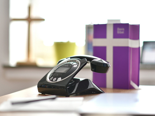 Schreibtisch mit Focus auf Telefon, die Basisbibel unscharf dahinter. Im Hintergrund in der Unschärfe, die Fensterbank mit Kreuz und Blumentopf.