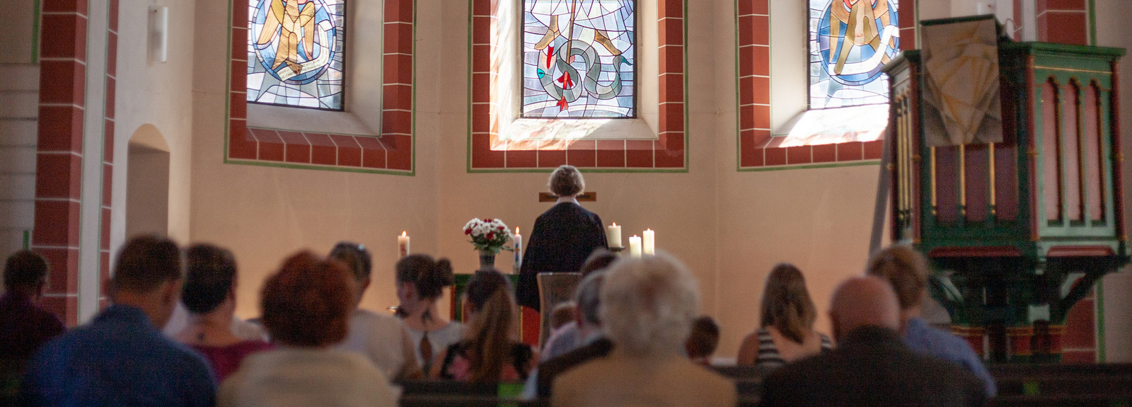 Evangelischer Gottesdienst in einer Kirche. Eine Pastorin wendet sich zum Altar. Die Gemeinde sitzt in den Bänken und richtet den Blick ebenfalls zum Altar. 