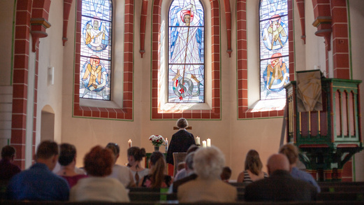 Evangelischer Gottesdienst in einer Kirche. Eine Pastorin wendet sich zum Altar. Die Gemeinde sitzt in den Bänken und richtet den Blick ebenfalls zum Altar. 