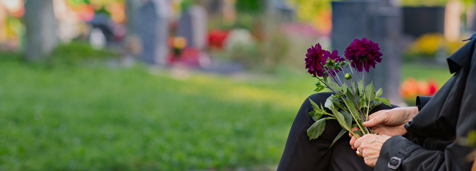 Ein Besucher eines Friedhofs sitzt und hält einige Blumen in der Hand. Im unscharfen Hintergrund sind Grabsteine zu erkennen.