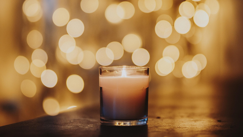 Eine Kerze in einem Glas steht mittig auf einem Holztisch. Im Hintergrund sind verschwommen viele Lichtpunkte zu sehen. 