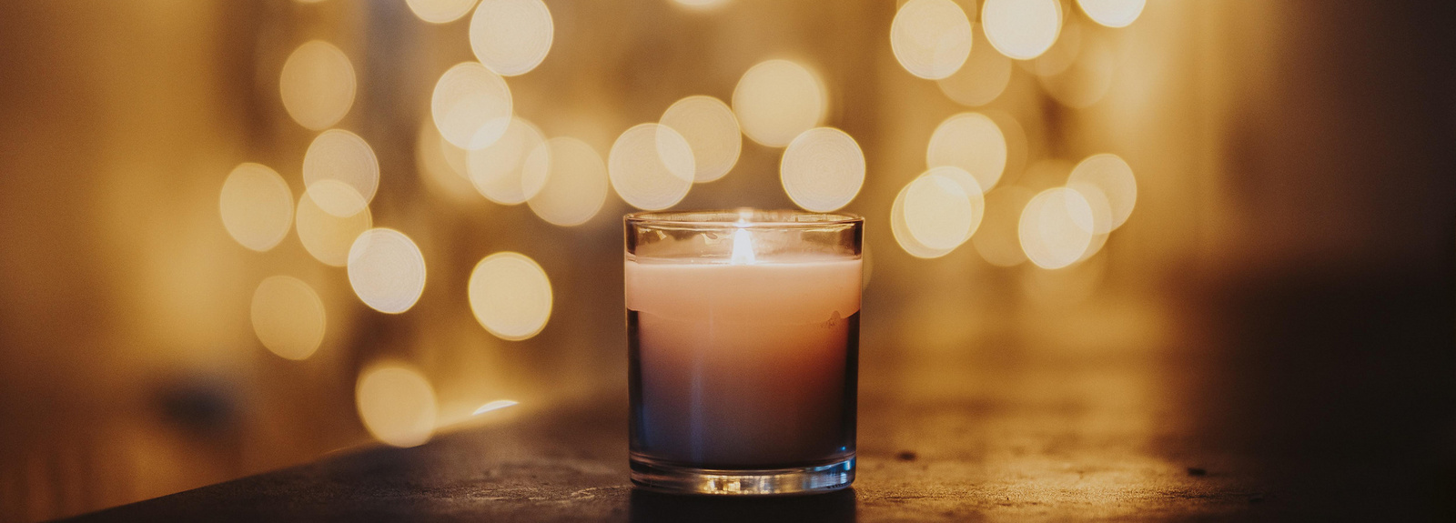 Eine Kerze in einem Glas steht mittig auf einem Holztisch. Im Hintergrund sind verschwommen viele Lichtpunkte zu sehen. 