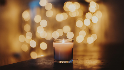 Eine Kerze in einem Glas steht mittig auf einem Holztisch. Im Hintergrund sind verschwommen viele Lichtpunkte zu sehen. 