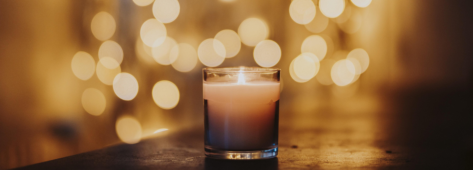 Eine Kerze in einem Glas steht mittig auf einem Holztisch. Im Hintergrund sind verschwommen viele Lichtpunkte zu sehen. 