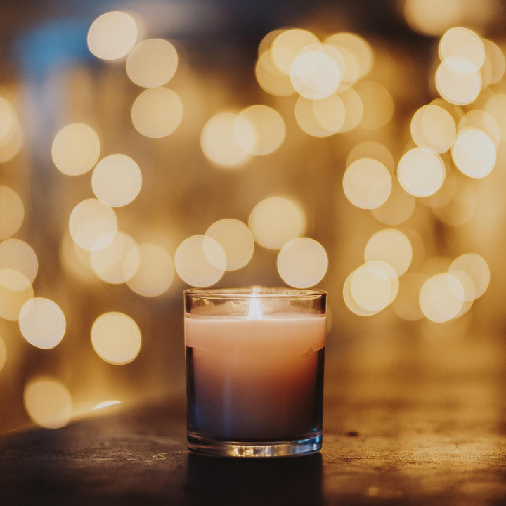 Eine Kerze in einem Glas steht mittig auf einem Holztisch. Im Hintergrund sind verschwommen viele Lichtpunkte zu sehen. 