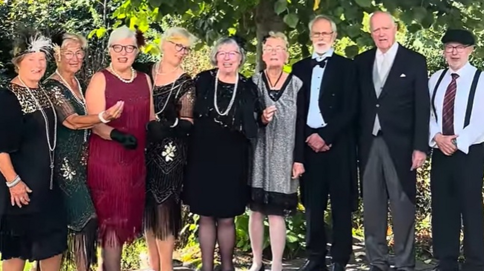 Eine Gruppe von Chormitgliedern in Kleidung der 20er Jahre des 20. Jahrhunderts steht vor einem grünen Hintergrund aus Bäumen und Sträuchern.