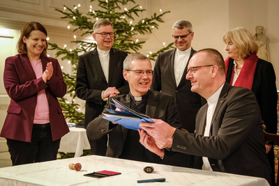 Bischof Dr. Heiner Wilmer, Bistum Hildesheim (links), und Bischof Thomas Adomeit, Ratsvorsitzender der Konföderation evangelischer Kirchen in Niedersachsen (rechts), tauschen die unterschriebene Vereinbarung zum neuen Unterrichtsfach Christliche Religion aus. Links daneben steht die niedersächsische Kultusministerin Julia Willie Hamburg. 