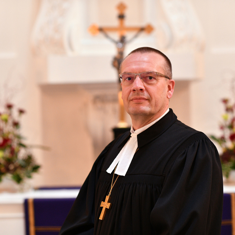 Bischof Thomas Adomeit im Talar schaut in die Kamera. Im hellen Hintergrund ist verschwommen ein Altar und ein Kreuz zu sehen.